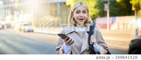Portrait of young blond woman with backpack, holding mobile phone, standing on street on sunny day, looking surprised and amazed at camera, expresses interest, hear great news 109641520