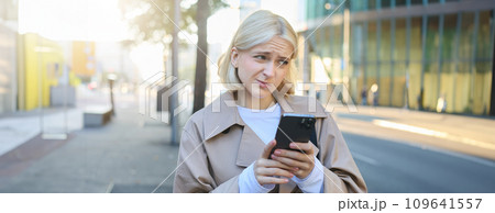 Portrait of young blond woman standing on street, has unsure, doubtful face expression, using mobile phone, showing reluctance Portrait of young blond woman standing on street, has unsure, doubtful face expression, using mobile phone, showing reluctance 109641557