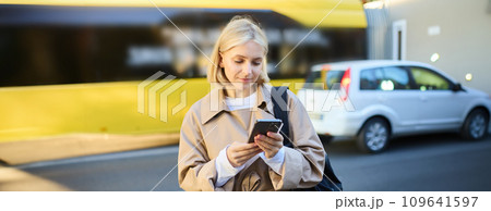 Outdoor shot of young woman with backpack, standing on street with cars passing behind her, holding mobile phone and smiling, urban lifestyle concept Outdoor shot of young woman with backpack, standing on street with cars passing behind her, holding mobile phone and smiling, urban lifestyle concept 109641597
