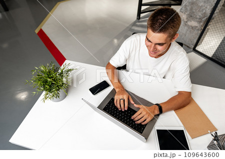 Casual young man in white t-shirt sitting at white desk looking at laptop screen at modern office Casual young man in white t-shirt sitting at white desk looking at laptop screen at modern office 109643600