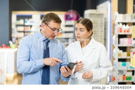 Middle-aged male customer asking young chemist about product reflected on his phone 109648004