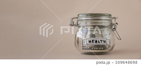 Saving Money In Glass Jar filled with Dollars banknotes. HEALTH transcription in front of jar. Managing personal finances extra income for future insecurity Saving Money In Glass Jar filled with Dollars banknotes. HEALTH transcription in front of jar. Managing personal finances extra income for future insecurity 109648698