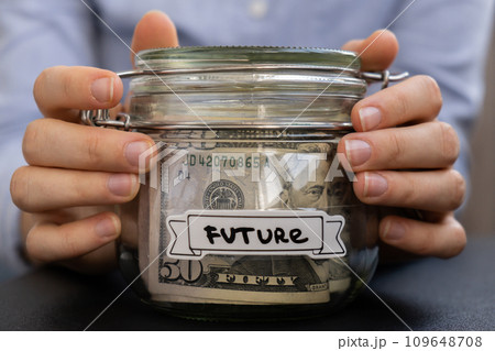 Unrecognizable woman holding Saving Money In Glass Jar filled with Dollars banknotes. FUTURE transcription in front of jar. Managing personal finances extra income for future insecurity 109648708