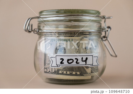 Saving Money In Glass Jar filled with Dollars banknotes. 2024 year transcription in front of jar. Managing personal finances extra income for future insecurity 109648710