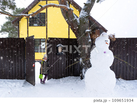 A child with a shovel at the gate and Funny snowman in a hat and scarf on the background of a yellow house in the yard. Winter, winter entertainment, snowfall 109651934