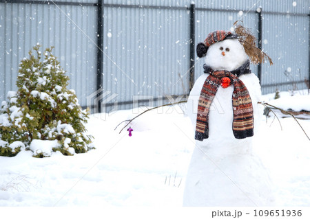 Funny snowman in a hat and scarf on the background of grey fence in the yard. Winter, winter entertainment, snowfall 109651936