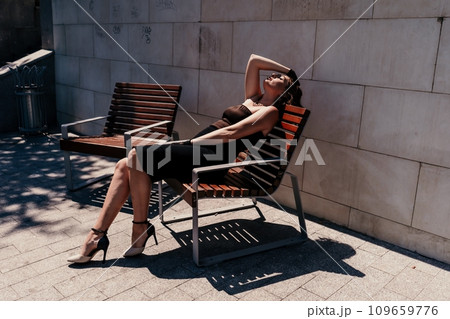 Portrait of a woman on the street. An attractive woman in a black dress is sitting on a bench outside. 109659776