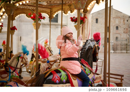 Happy child girl smiles and waves hello with hand while having fun on a merry go round carousel at Christmas funfair. Happy child girl smiles and waves hello with hand while having fun on a merry go round carousel at Christmas funfair. 109661698