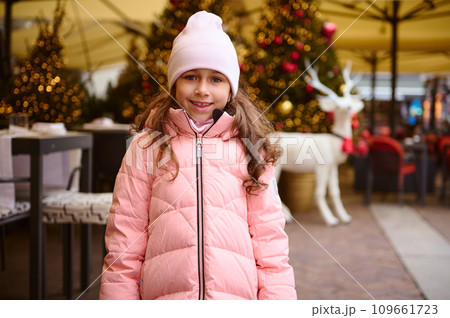 Lovely happy little kid girl smiles at camera, standing outdoors at Christmas fair, enjoying the upcoming festive events 109661723