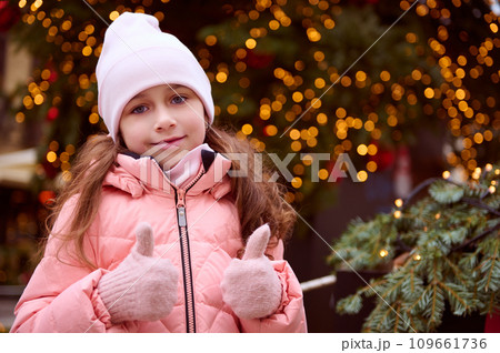 Cute little girl having fun at Christmas fair, showing thumbs up at camera, standing by illuminated Christmas tree 109661736