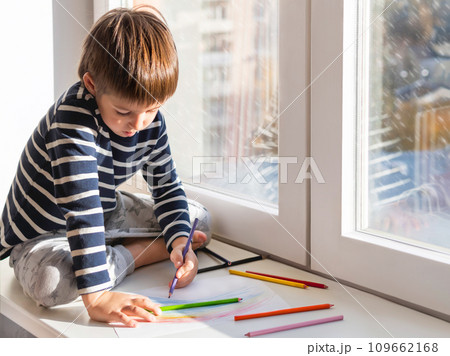 Left-handed boy sits on window sill and draws rainbow with colored pencils. Creative leisure activity for children at home. 109662168