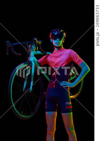 Young man in sportswear, helmet and googles holding bicycle on shoulder isolated over black studio background in neon light 109663733