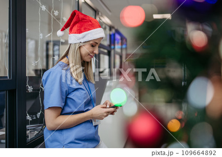 Portrait of beautiful nurse with christmas hat in hospital corridor, holding smartphone. Working in hospital on Christmas day, Christmas Eve. Female doctor working a Christmas shift and can't be with Portrait of beautiful nurse with christmas hat in hospital corridor, holding smartphone. Working in hospital on Christmas day, Christmas Eve. Female doctor working a Christmas shift and can't be with 109664892