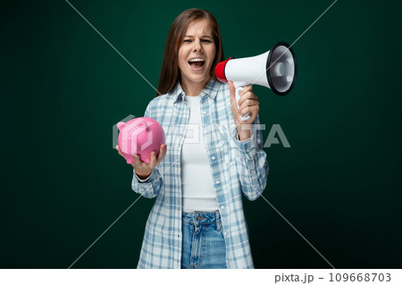 A cute young woman in a plaid shirt talks about financial literacy while holding a piggy bank in her A cute young woman in a plaid shirt talks about financial literacy while holding a piggy bank in her 109668703