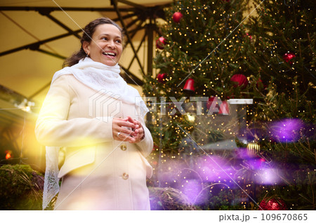 smiling woman at funfair, looking away, standing against a Christmas tree decorated with garlands. Bokeh on foreground 109670865