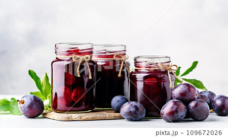 Ripe plums and plum compote in glass jars on the table. Home canned food and harvest of appetizing plums on a white background. Ripe plums and plum compote in glass jars on the table. Home canned food and harvest of appetizing plums on a white background. 109672026