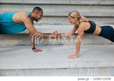 Multiracial sportive woman and man holding hands while standing in plank outdoor. 109672896
