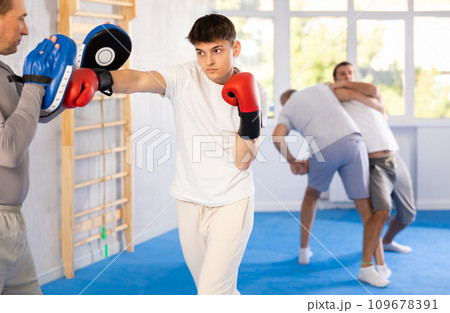 Coach holding mitts during self defence training in gym with young guy mastering base punches in boxing gloves in gym 109678391