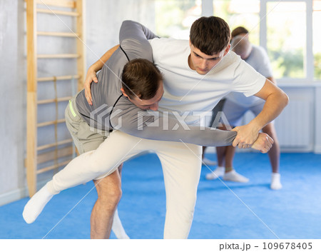 Concentrated man and guy in sport club during self-defense training, sparring and practicing technique of hip flip. In background, group of men practice self-defense techniques 109678405