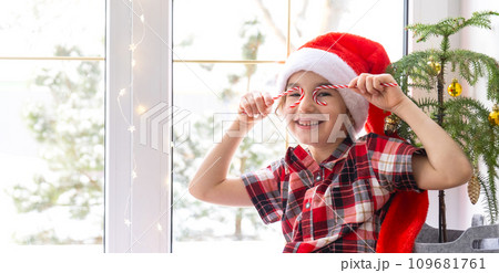 Girl in a Santa hat sits on the windowsill of a house near the Christmas tree and puts Candy cane to her eyes like glasses. Child is having fun and making faces, waiting for Christmas and New year Girl in a Santa hat sits on the windowsill of a house near the Christmas tree and puts Candy cane to her eyes like glasses. Child is having fun and making faces, waiting for Christmas and New year 109681761