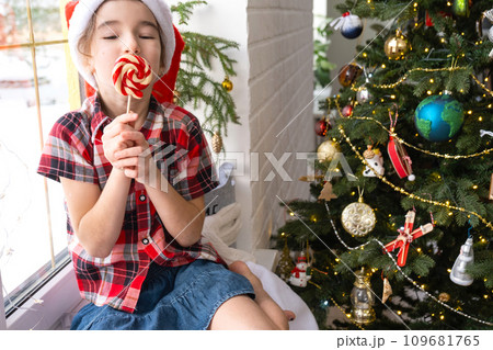 Happy Girl in a Santa hat sits on the windowsill of a house near the Christmas tree and puts sweet round caramel on a stick. Child is having fun and making faces, waiting for Christmas and New year Happy Girl in a Santa hat sits on the windowsill of a house near the Christmas tree and puts sweet round caramel on a stick. Child is having fun and making faces, waiting for Christmas and New year 109681765