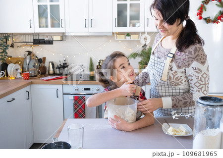 Mom and daughter in the white kitchen are preparing cookies, add ingredients. Family day, preparation for the holiday Christmas, learn to cook delicious pastries, cut shapes out of dough with molds Mom and daughter in the white kitchen are preparing cookies, add ingredients. Family day, preparation for the holiday Christmas, learn to cook delicious pastries, cut shapes out of dough with molds 109681965