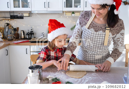 Mom and daughter in the white kitchen are preparing cookies for Christmas and new year. Family day, preparation for the holiday, learn to cook delicious pastries, cut shapes out of dough with molds 109682012