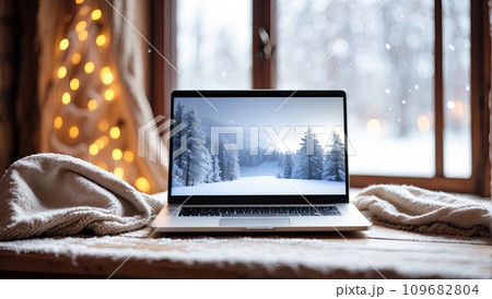 Laptop with a winter landscape screen, indoor on the table with a cozy blanket by the window with snow. Seasonal remote work, internet, shopping, Christmas and New Year. AI generated 109682804