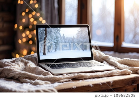 Laptop with a winter landscape screen, indoor on the table with a cozy blanket by the window with snow. Seasonal remote work, internet, shopping, Christmas and New Year. AI generated 109682808