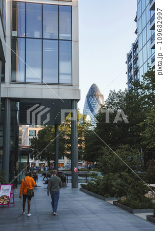 Tourists walking on sidewalk with The Gherkin in background. Office buildings with blue sky in background. View of financial district in London. 109682997