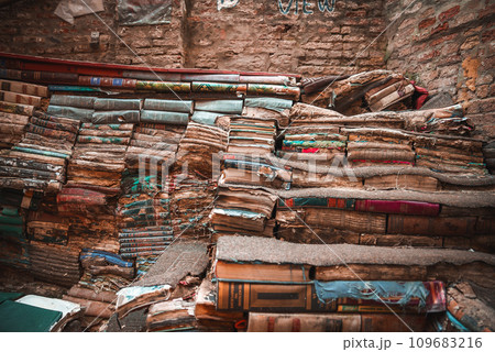 A dimly lit room in an old, weathered building in Venice. Sunlight streams in, casting a warm glow on a haphazard pile of old books, creating a cozy and inviting space. 109683216