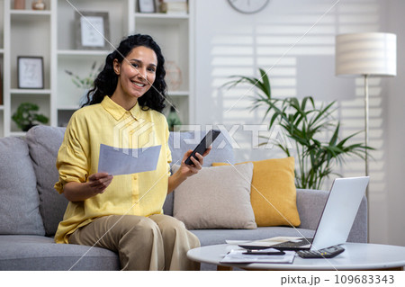Satisfied businesswoman working from home, sitting on sofa in front of laptop, holding phone and documents, smiling and looking at camera. Satisfied businesswoman working from home, sitting on sofa in front of laptop, holding phone and documents, smiling and looking at camera. 109683343