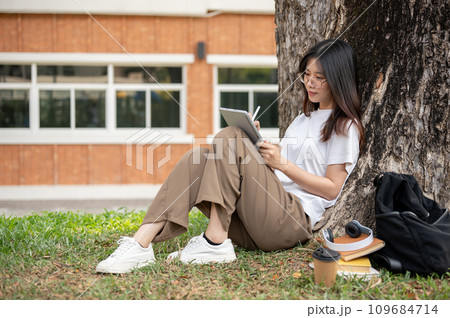 A beautiful Asian female student is using her digital tablet under the tree in a campus park. A beautiful Asian female student is using her digital tablet under the tree in a campus park. 109684714