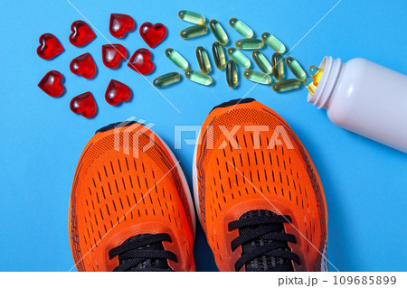 A jar of scattered omega 3 capsules and decorative hearts in front of orange sports sneakers on a blue background A jar of scattered omega 3 capsules and decorative hearts in front of orange sports sneakers on a blue background 109685899