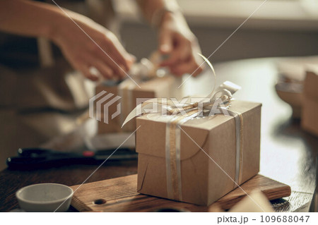 A craft box with white and gold ribbons. A woman's hands ties a ribbon on a customer's order box. A small business entrepreneur and the concept of food delivery. 109688047
