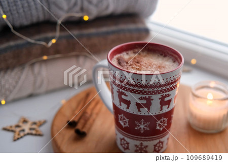 Winter windowsill still life. Red ceramic cup of hot coffee on window sill. Christmas decorations on the background. Cozy home picture. Warm woolen knitted sweaters, Burn Candle, Cookies. Stock photo 109689419