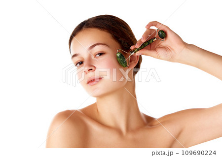 Beautiful young girl taking care after skin, using face massage roller against white background. Face lifting, refreshment Beautiful young girl taking care after skin, using face massage roller against white background. Face lifting, refreshment 109690224