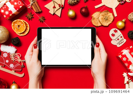 Top view of woman holding tablet in her hands on red background made of Christmas decorations. New Year holiday concept. Mockup Top view of woman holding tablet in her hands on red background made of Christmas decorations. New Year holiday concept. Mockup 109691464
