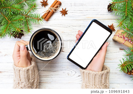 Female hands holding modern smartphone with mosk up and mug of coffee on wooden table with christmas decoration. top view Female hands holding modern smartphone with mosk up and mug of coffee on wooden table with christmas decoration. top view 109691843