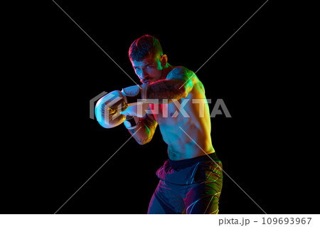 Young man, shirtless boxing athlete with muscular body training, punching isolated over black background in neon light Young man, shirtless boxing athlete with muscular body training, punching isolated over black background in neon light 109693967