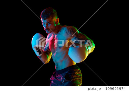 Competitive, concentrated strong, muscular young man, boxing athlete training shirtless isolated over black background in neon light Competitive, concentrated strong, muscular young man, boxing athlete training shirtless isolated over black background in neon light 109693974