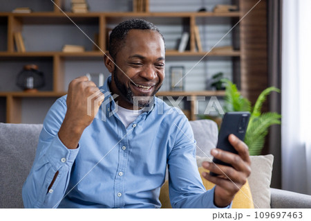 Online winner man sitting at home on sofa in living room, happy successful african american man celebrating victory, holding hand up, received online notification on phone, happy and satisfied. Online winner man sitting at home on sofa in living room, happy successful african american man celebrating victory, holding hand up, received online notification on phone, happy and satisfied. 109697463