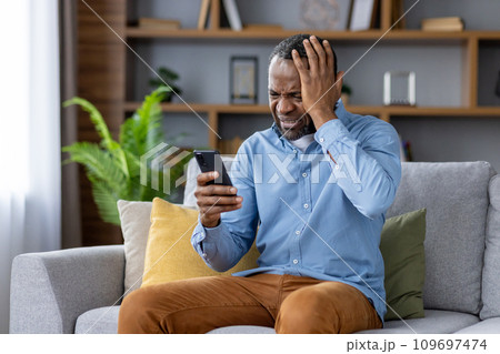 Sad nervous man sitting at home on sofa in living room of house, African American man received bad news online, read news on social media, received negative feedback via email. Sad nervous man sitting at home on sofa in living room of house, African American man received bad news online, read news on social media, received negative feedback via email. 109697474
