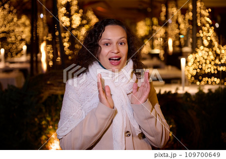 Beautiful woman expressing amazement standing against a city street illuminated with Christmas garlands at night time Beautiful woman expressing amazement standing against a city street illuminated with Christmas garlands at night time 109700649