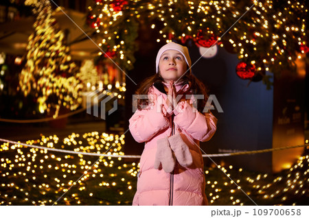 Adorable child girl holding hands palms together, making cherished wish, standing against a Christmas tree at funfair 109700658