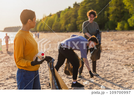 Earth day. Volunteers activists team collects garbage cleaning of beach coastal zone. Woman puts plastic bottle trash in garbage bag on ocean shore. Environmental conservation coastal zone cleaning 109700997