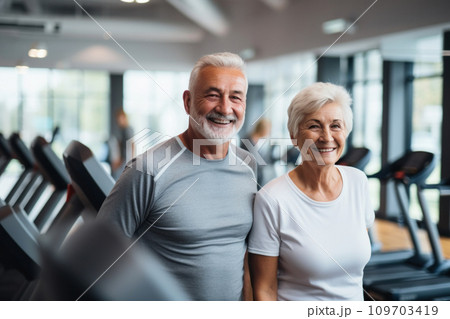 Cheerful smiling senior couple in grey sports attire in modern gym with gym's equipment in background,, active and healthy lifestyle and joy of shared activities, fitness and well-being at any age. 109703419