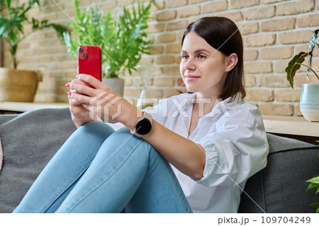 Young happy woman using smartphone, sitting on sofa at home Young happy woman using smartphone, sitting on sofa at home 109704249