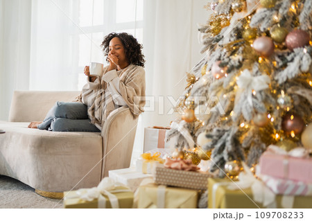 Merry Christmas. African American woman with cup of hot drink coffee tea sitting near Christmas tree at home. Girl in living room with Christmas tree. Christmas hygge holidays at home 109708233