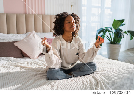 Yoga mindfulness meditation. Young healthy African girl practicing yoga at home. Woman sitting in lotus pose on bed meditating smiling relaxing indoor. Girl doing breathing practice. Yoga at home Yoga mindfulness meditation. Young healthy African girl practicing yoga at home. Woman sitting in lotus pose on bed meditating smiling relaxing indoor. Girl doing breathing practice. Yoga at home 109708234